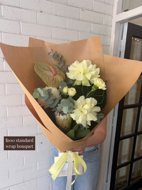 Bouquet of flowers wrapped in brown paper held by a person against a white brick wall.