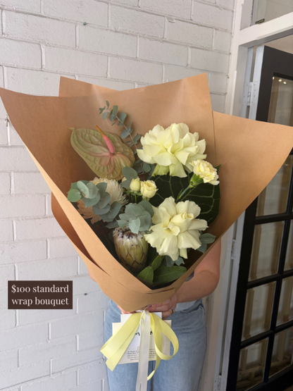 Bouquet of flowers wrapped in brown paper held by a person against a white brick wall.
