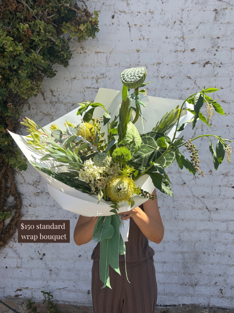 Person holding a large bouquet of greenery against a white brick wall.