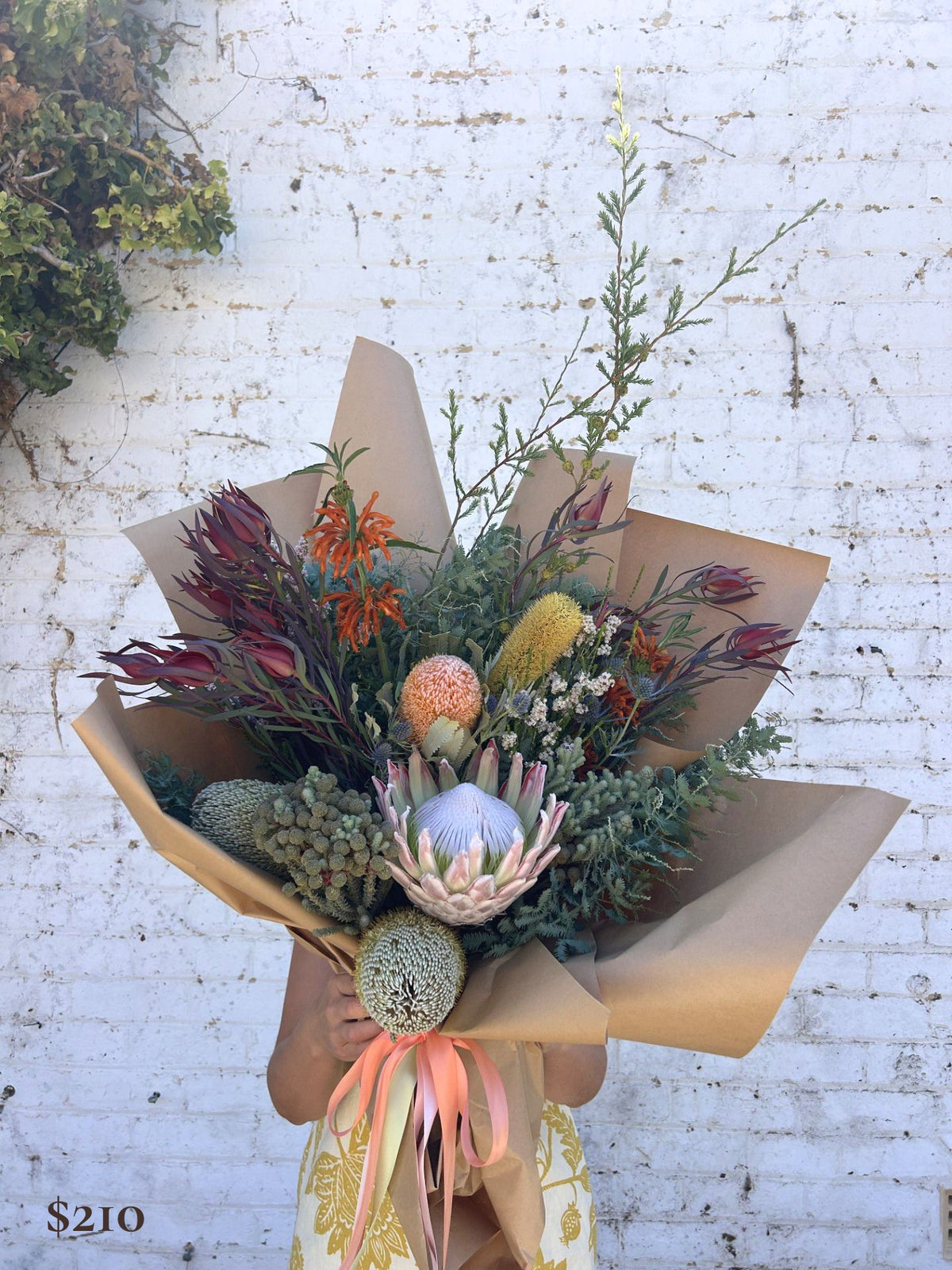 A hand-held dried flower bouquet wrapped in brown kraft paper and tied with a peach ribbon, featuring a variety of earthy botanicals against a white brick wall background.