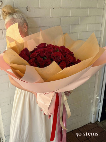 A large bouquet of 50 deep red roses wrapped in beige and light pink paper with red and pink ribbons, held by a woman in a white dress against a white brick wall.