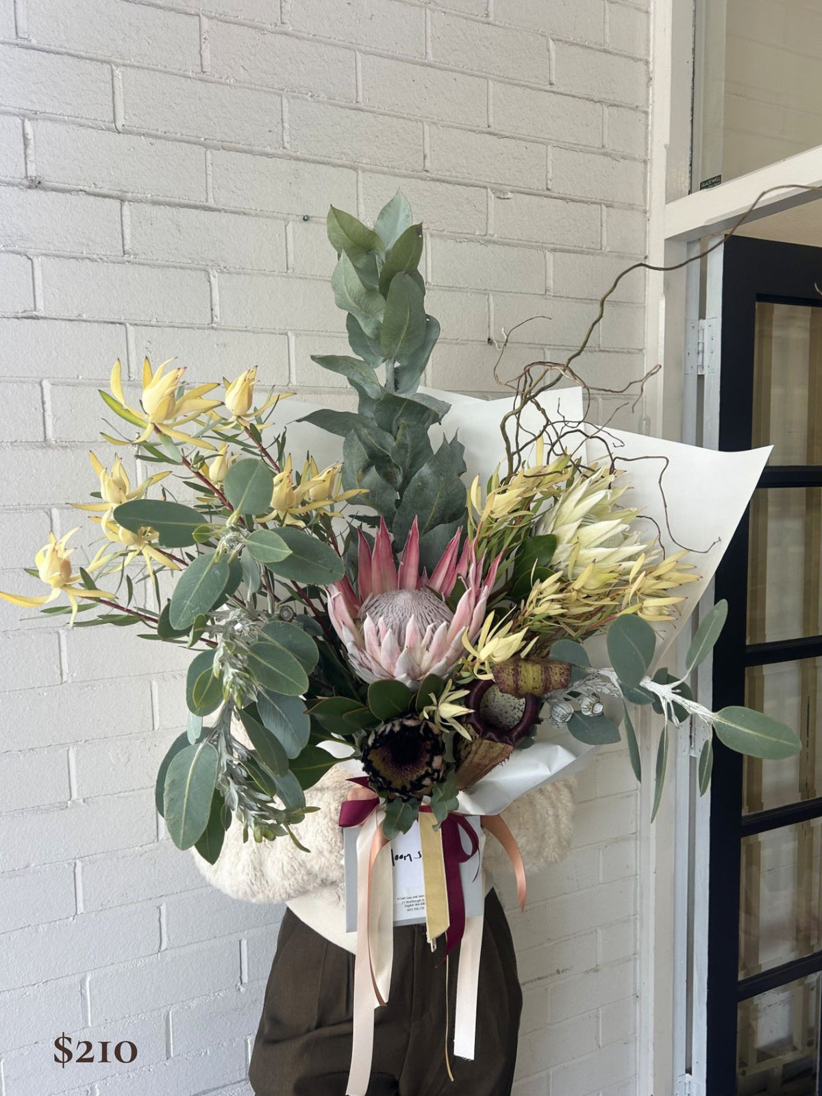 A large, beautifully arranged dried and live floral arrangement featuring a prominent pink protea, yellow flowers, green eucalyptus, and brown botanical elements, wrapped in white paper and tied with cream and burgundy ribbons, held in front of a white brick wall.