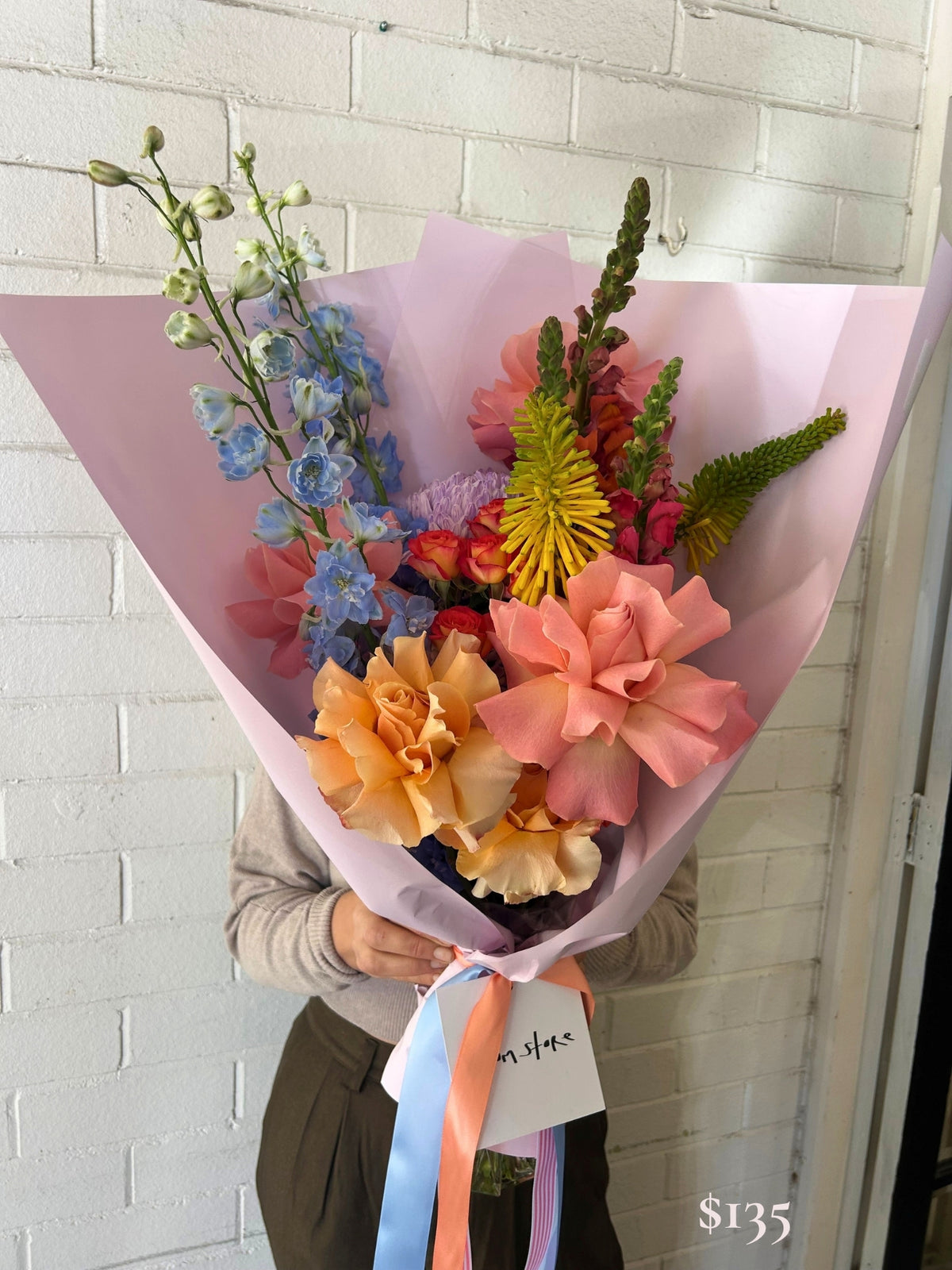 A person holds a large, vibrant floral bouquet wrapped in light pink paper, featuring peach roses, blue delphiniums, yellow chrysanthemums, and green foliage, set against a white brick wall.