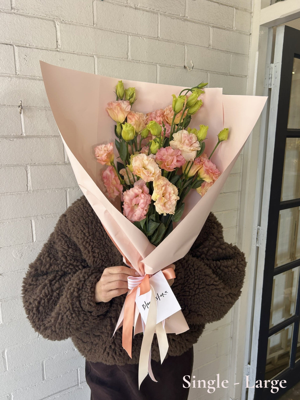A person wearing a brown sherpa fleece jacket holds a large bouquet of pink and peach lisianthus flowers wrapped in pale blush paper with a ribbon and a small card, standing against a white painted brick wall.