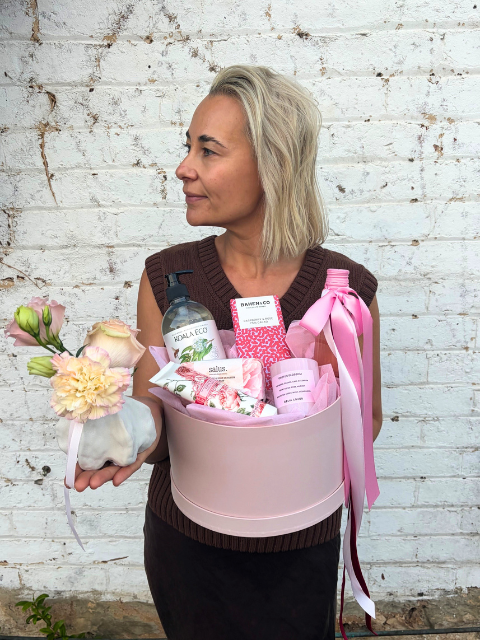 Woman holding a gift basket with various items against a white brick wall.