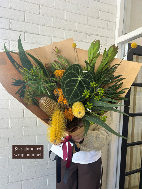 Person holding a large bouquet of flowers wrapped in brown paper against a white brick wall.