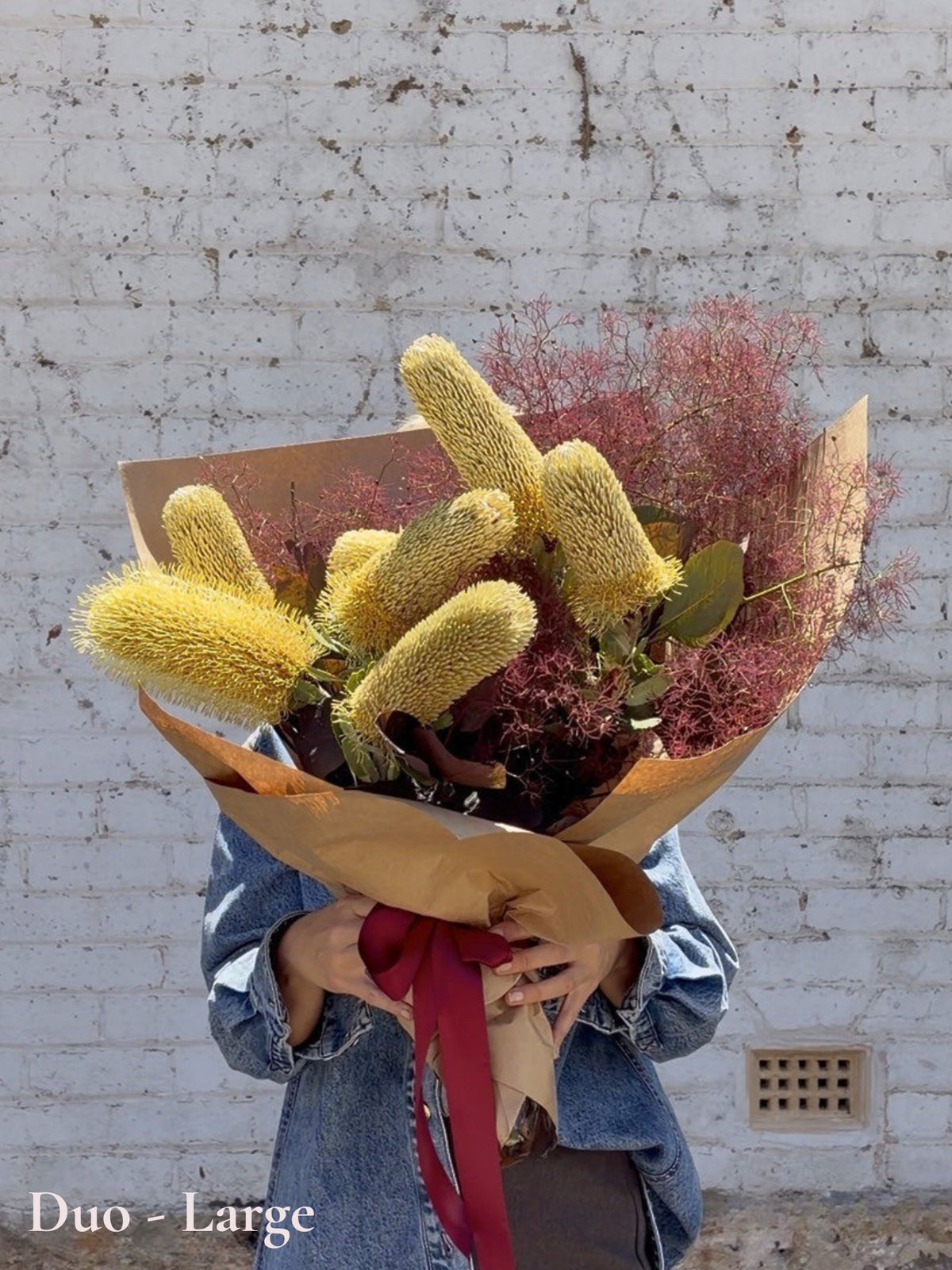 A person in a denim jacket holds a large dried flower bouquet wrapped in brown paper with a red ribbon, set against a white brick wall. The bouquet features prominent golden-yellow banksia and dusty pink/purple smokebush.