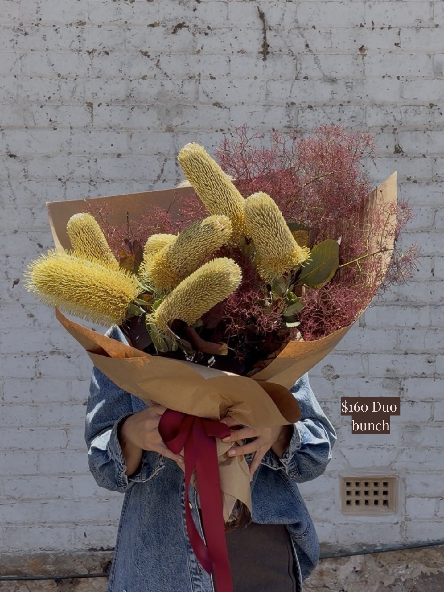 A person holding a native flower bouquet against a white brick wall.