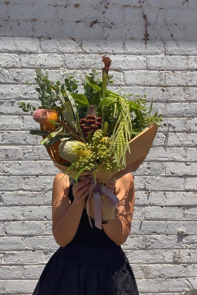 Person holding a bouquet of greenery and natives against a gray brick wall