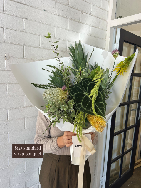 Person holding a bouquet of flowers wrapped in paper against a white brick wall.