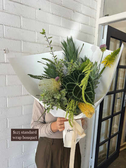 Person holding a bouquet of flowers wrapped in paper against a white brick wall.