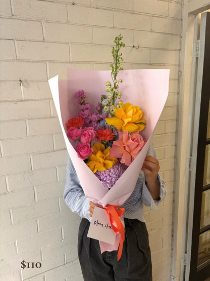 A person holding a vibrant multicolor flower bouquet wrapped in light pink paper with a red ribbon and a 'Flower Shop' card, standing against a white painted brick wall.