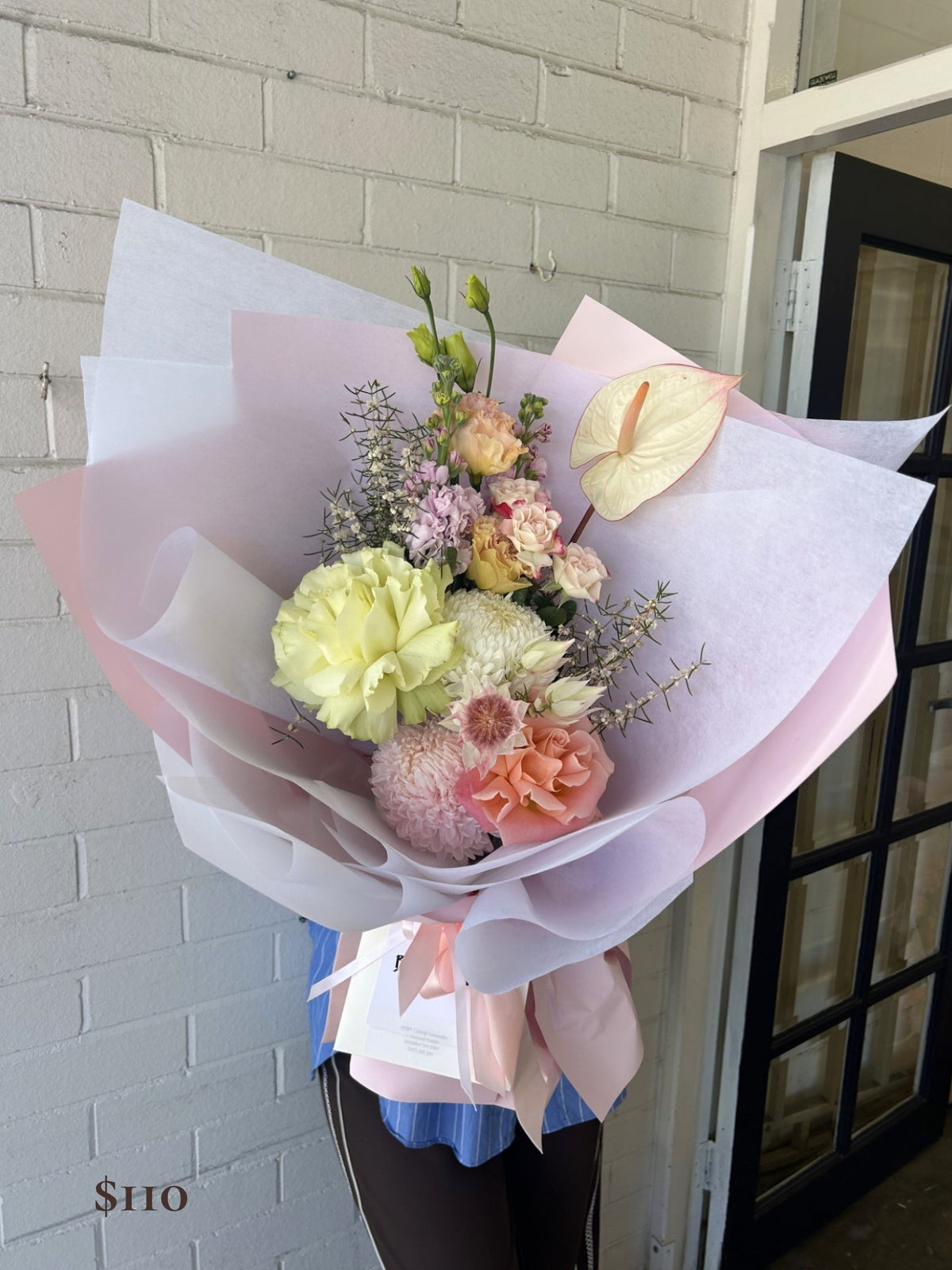 A hand-held flower bouquet wrapped in pale pink and white tissue paper, featuring pastel yellow, peach, pink, and cream flowers with green stems, held in front of a white brick wall and glass door.
