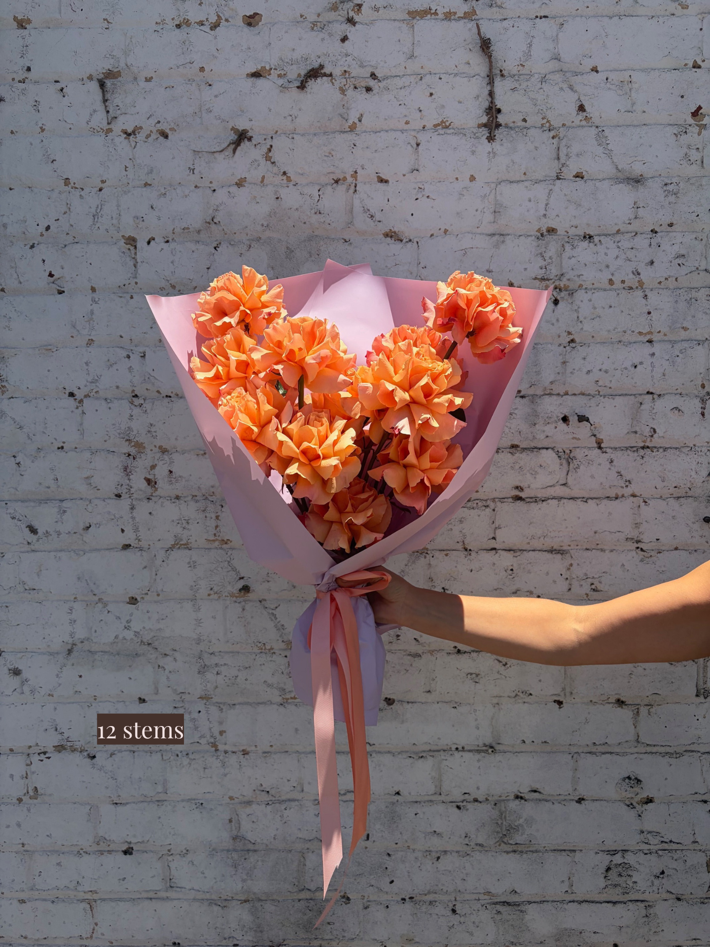 Bouquet of orange flowers with pink paper and ribbon held against a white brick wall.