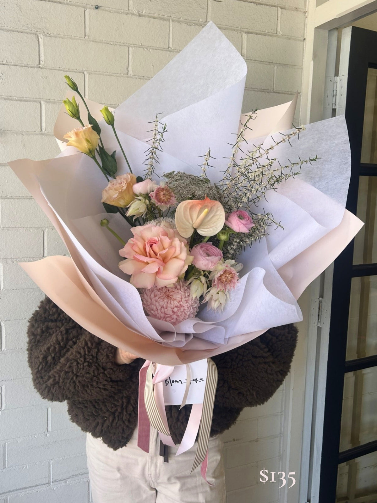 A hand-held flower bouquet wrapped in white and blush pink paper, featuring pink roses, peach blossoms, anthuriums, and green filler stems, held by someone wearing a dark brown fuzzy sweater in front of a white brick wall.