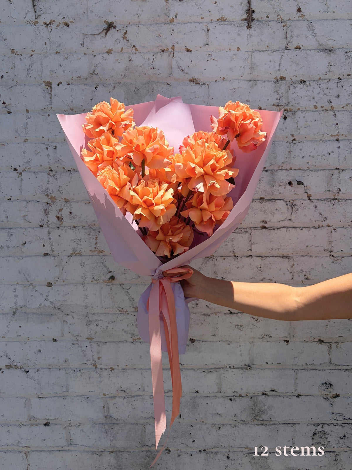 A bouquet of 12 orange artificial flowers wrapped in light pink paper and tied with a pink ribbon, held by a person's hand against a white brick wall background.