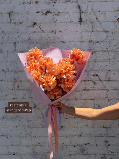 Bouquet of orange flowers with a pink wrap held against a textured white wall.