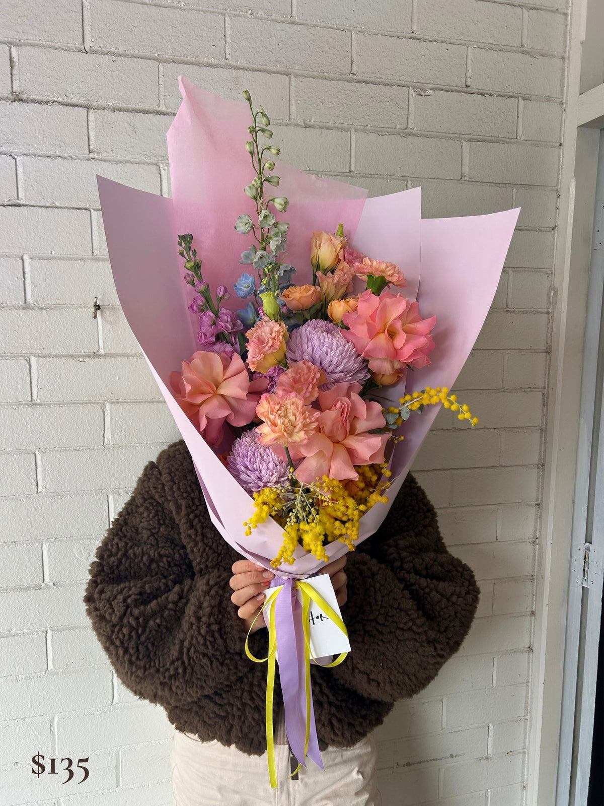 A person wearing a brown sherpa jacket holds a large bouquet of pink, purple, and yellow flowers wrapped in light pink paper, standing against a white brick wall.