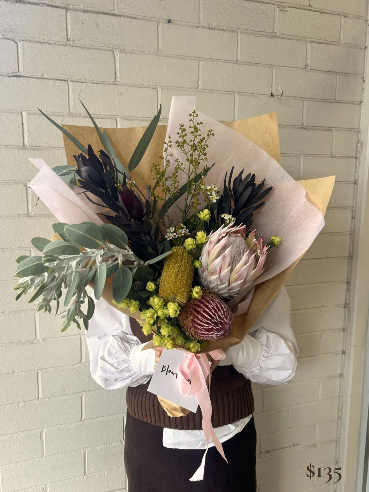 A person holding a large dried and wildflower bouquet wrapped in kraft paper and blush pink tissue. The bouquet features green eucalyptus, dark burgundy protea-like flowers, pale pink protea, yellow-green bursaria, and small white flowers, with a pink ribbon and a handwritten price tag.