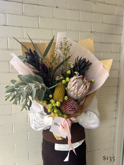 A person holding a large dried and wildflower bouquet wrapped in kraft paper and blush pink tissue. The bouquet features green eucalyptus, dark burgundy protea-like flowers, pale pink protea, yellow-green bursaria, and small white flowers, with a pink ribbon and a handwritten price tag.