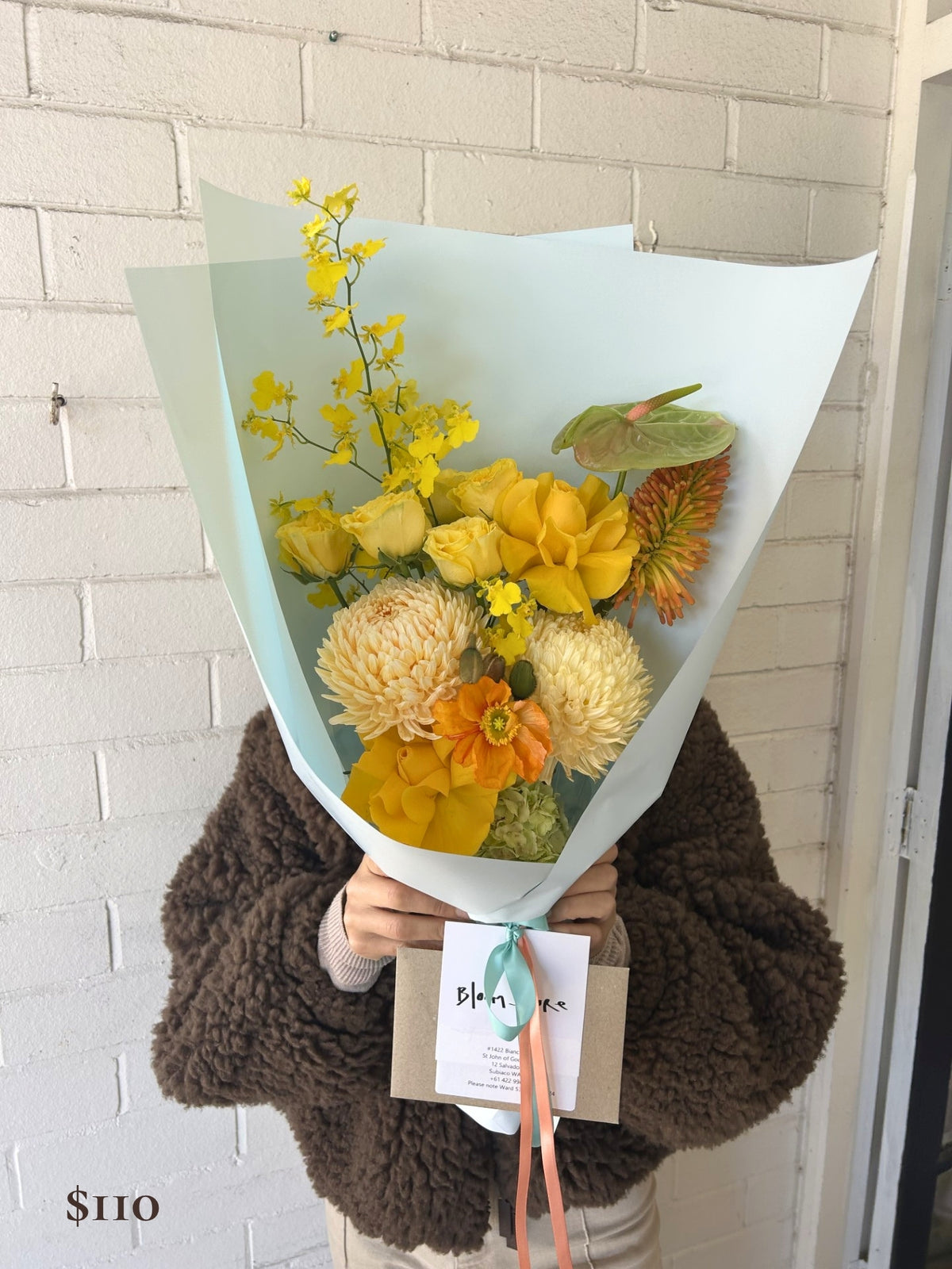 A person in a brown fuzzy sweater holds a wrapped bouquet of yellow and orange flowers in front of a white brick wall. The bouquet is wrapped in pale blue paper with a small card tied at the bottom.