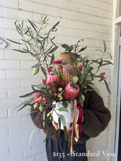 A person in a brown fuzzy jacket holds a large, lush floral arrangement featuring pink protea-like flowers, green eucalyptus leaves, and olive green pods in a white pot, set against a white painted brick wall.