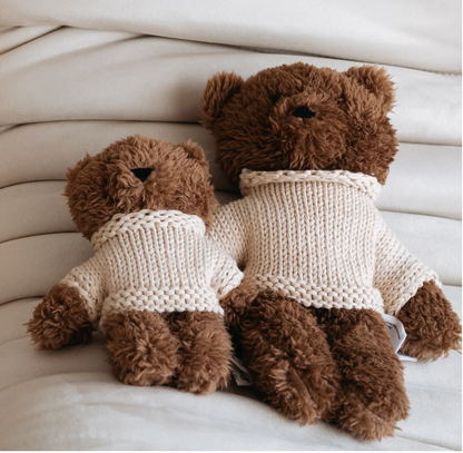 Two brown teddy bears wearing matching beige sweaters on a light-colored surface.