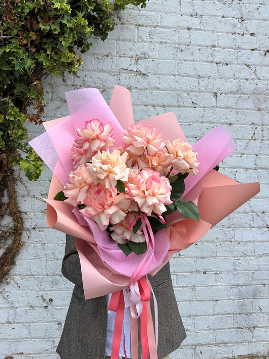 A bouquet of pink and peach carnations wrapped in pink tissue paper and peach paper, tied with pink and white ribbons, held by a person wearing a gray blazer against a white brick wall.