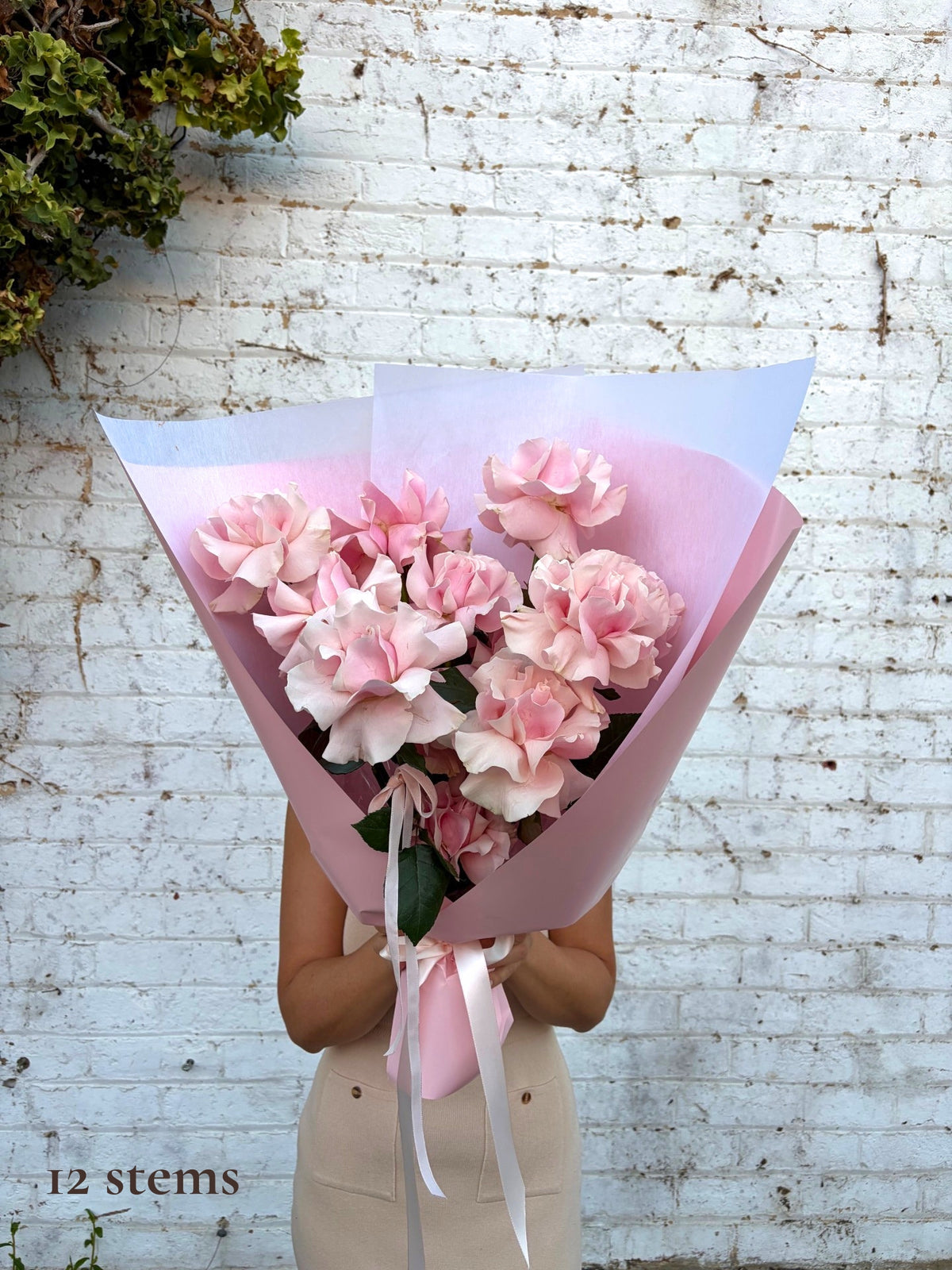 A person holding a bouquet of 12 pink roses wrapped in light pink and white paper, standing in front of a white brick wall.