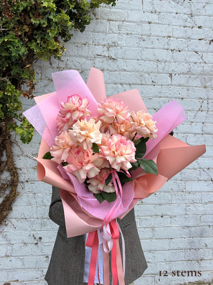 A bouquet of pink and peach carnations wrapped in pink tissue paper and peach paper, tied with pink and white ribbons, held by a person wearing a gray blazer against a white brick wall.