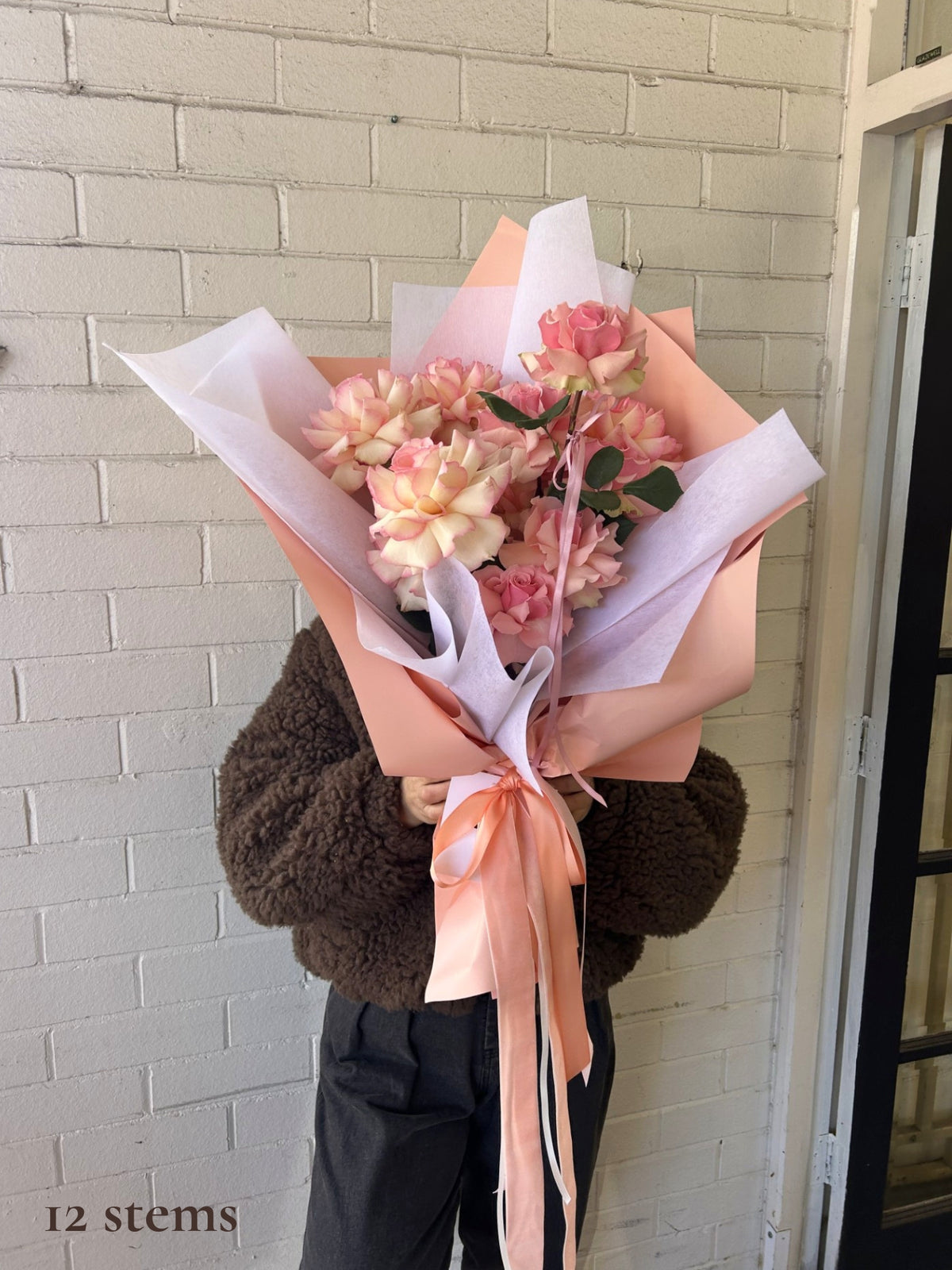 A person wearing a brown fuzzy jacket holds a large bouquet of pink and peach carnations wrapped in white and peach paper with a peach ribbon, standing against a white brick wall.