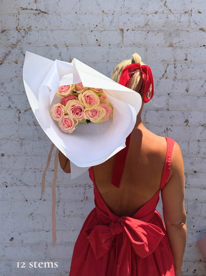 A woman seen from behind wearing a red backless dress with a large bow, holding a large bouquet of pink and yellow roses wrapped in white paper in front of her face. She stands against a white brick wall.