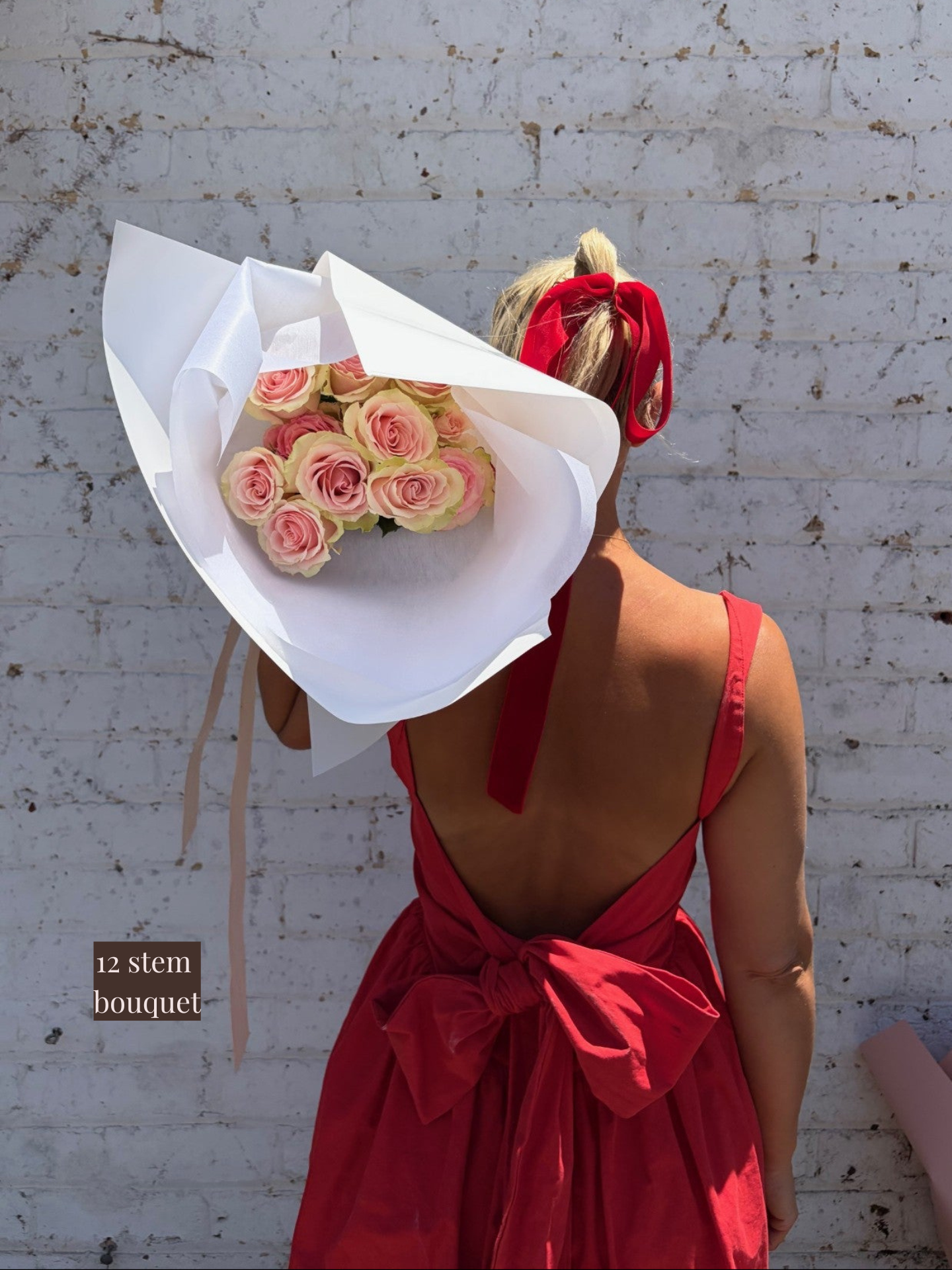 Person wearing a red ribbon bow with a bouquet of pink roses, standing against a textured white wall.