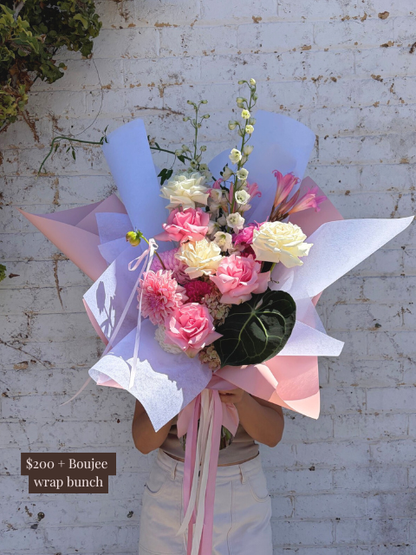 Bouquet of flowers wrapped in pink and white paper with a person holding it against a white brick wall.