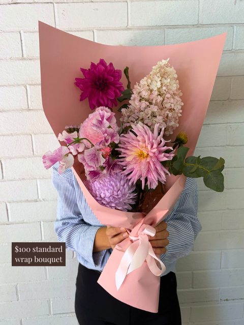 Bouquet of flowers wrapped in pink paper held by a person against a white brick wall.