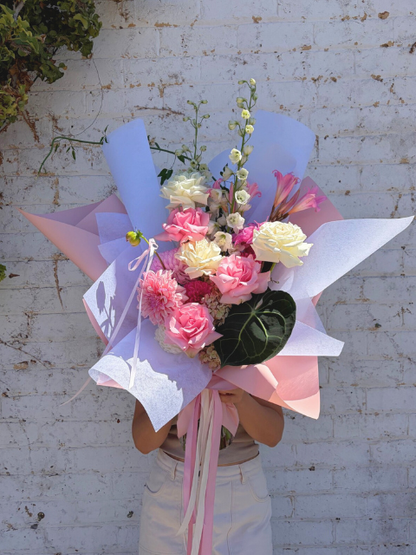 Bouquet of flowers wrapped in pink and white paper with a person holding it against a white brick wall.