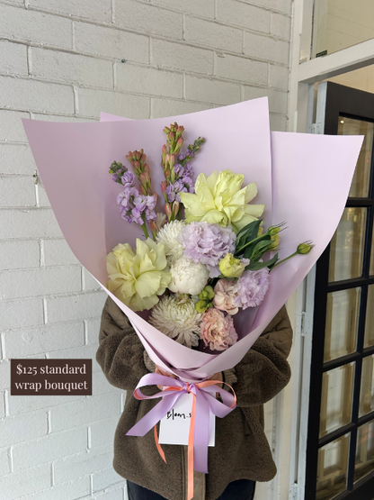 Bouquet of flowers wrapped in purple paper with a person holding it, against a light brick wall.