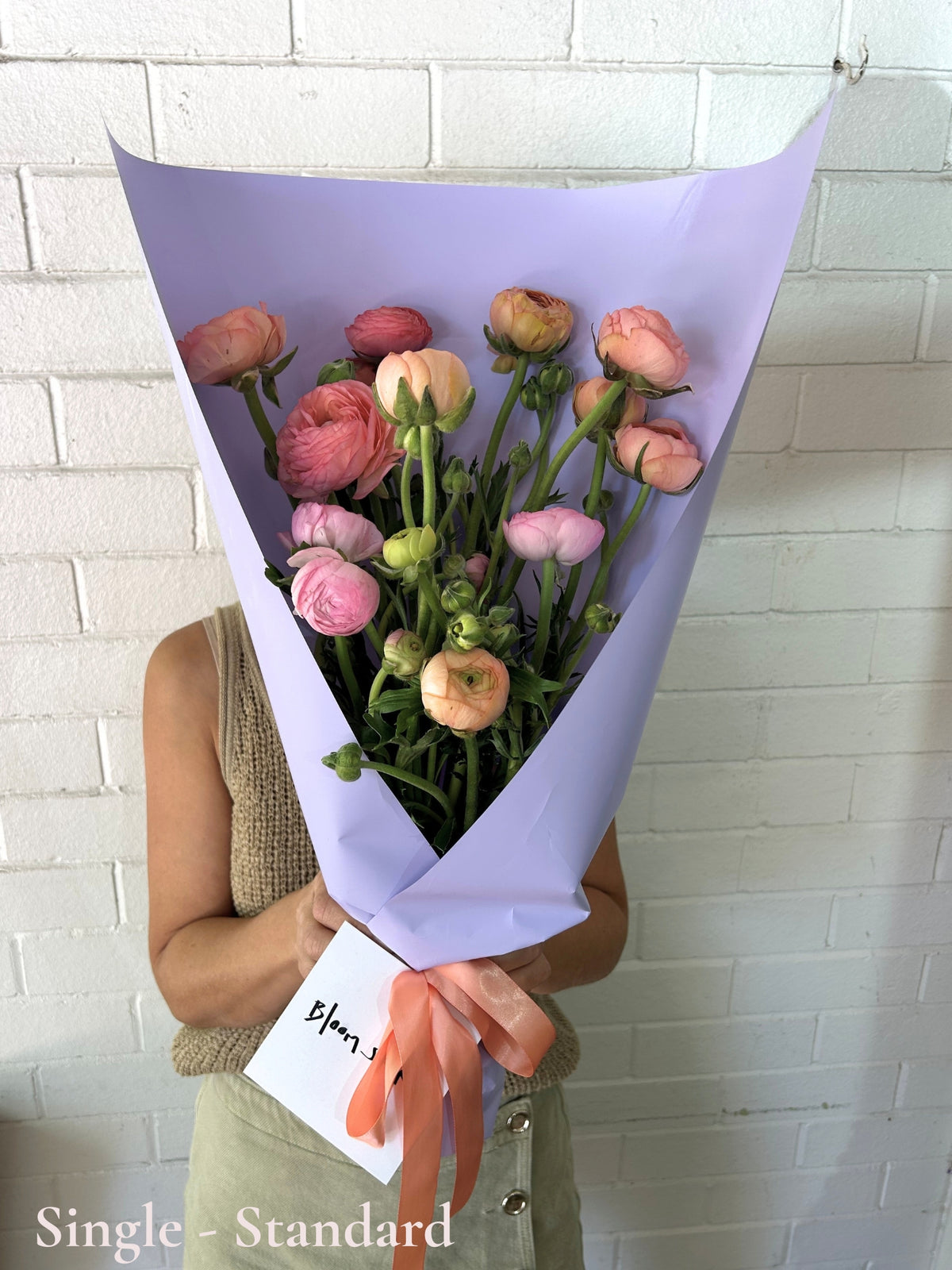 A bouquet of pink and peach ranunculus flowers wrapped in light purple paper with a peach ribbon and a small white card, held by a person standing against a white brick wall.