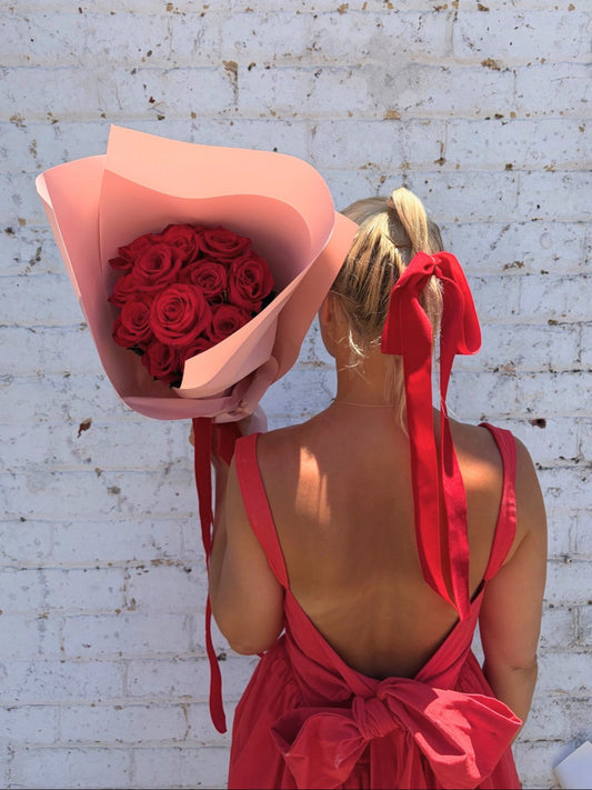 A woman with blonde hair in a ponytail wearing a red dress, seen from behind, holding a large bouquet of red roses wrapped in pink paper against a white brick wall.