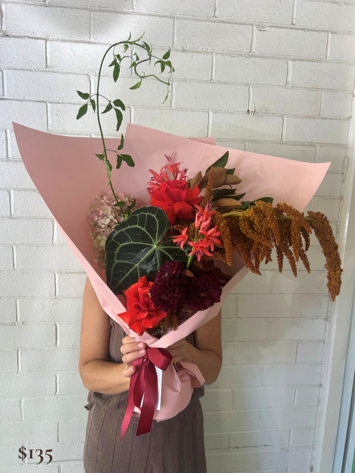 A person holds a large floral bouquet wrapped in light pink paper, featuring red and burgundy flowers, green leaves, and brown dried stems, in front of a white brick wall.