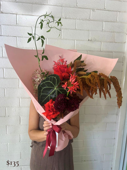 A person holds a large floral bouquet wrapped in light pink paper, featuring red and burgundy flowers, green leaves, and brown dried stems, in front of a white brick wall.