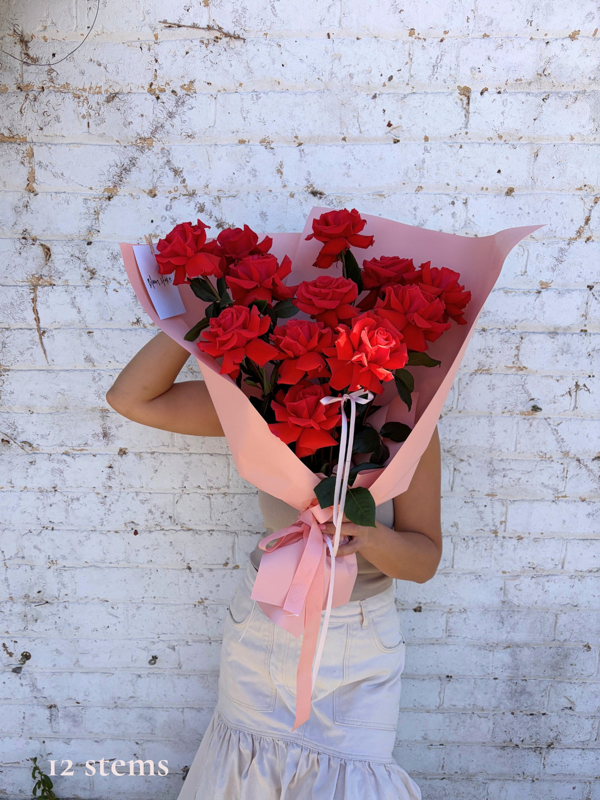 A person holding a large bouquet of 12 red roses wrapped in pink paper and tied with a pink ribbon, standing against a white brick wall.
