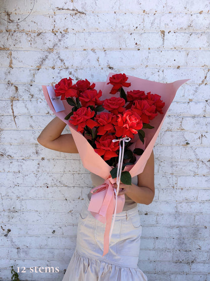 A person holding a large bouquet of 12 red roses wrapped in pink paper and tied with a pink ribbon, standing against a white brick wall.