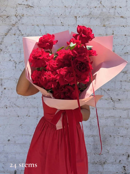 A bouquet of 24 red roses wrapped in light pink paper and a red ribbon, held by a person wearing a red dress against a white brick wall.