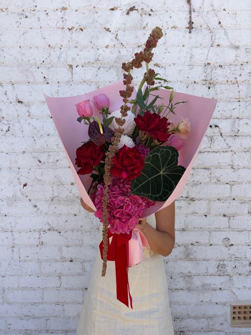 Person holding a bouquet of red and pink flowers against a white brick wall