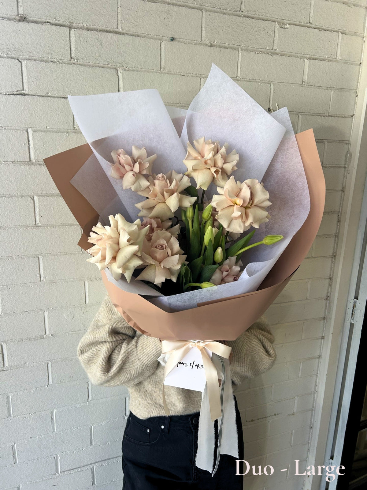 A bouquet of large pale peach  roses wrapped in brown kraft paper and white tissue paper, tied with a cream ribbon, held by a person wearing a beige sweater and dark jeans against a white painted brick wall.