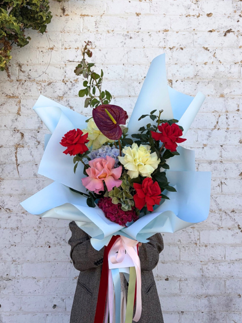 Bouquet of red, green, pink and blue flowers with blue wrapping against a textured white wall.