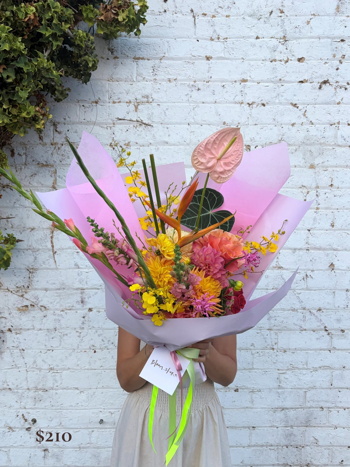 A person holding a large, vibrant floral bouquet wrapped in pink paper, standing against a white brick wall. The bouquet features a variety of colorful flowers including yellow, pink, orange, and green.