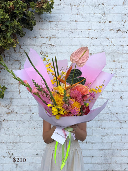 A person holding a large, vibrant floral bouquet wrapped in pink paper, standing against a white brick wall. The bouquet features a variety of colorful flowers including yellow, pink, orange, and green.