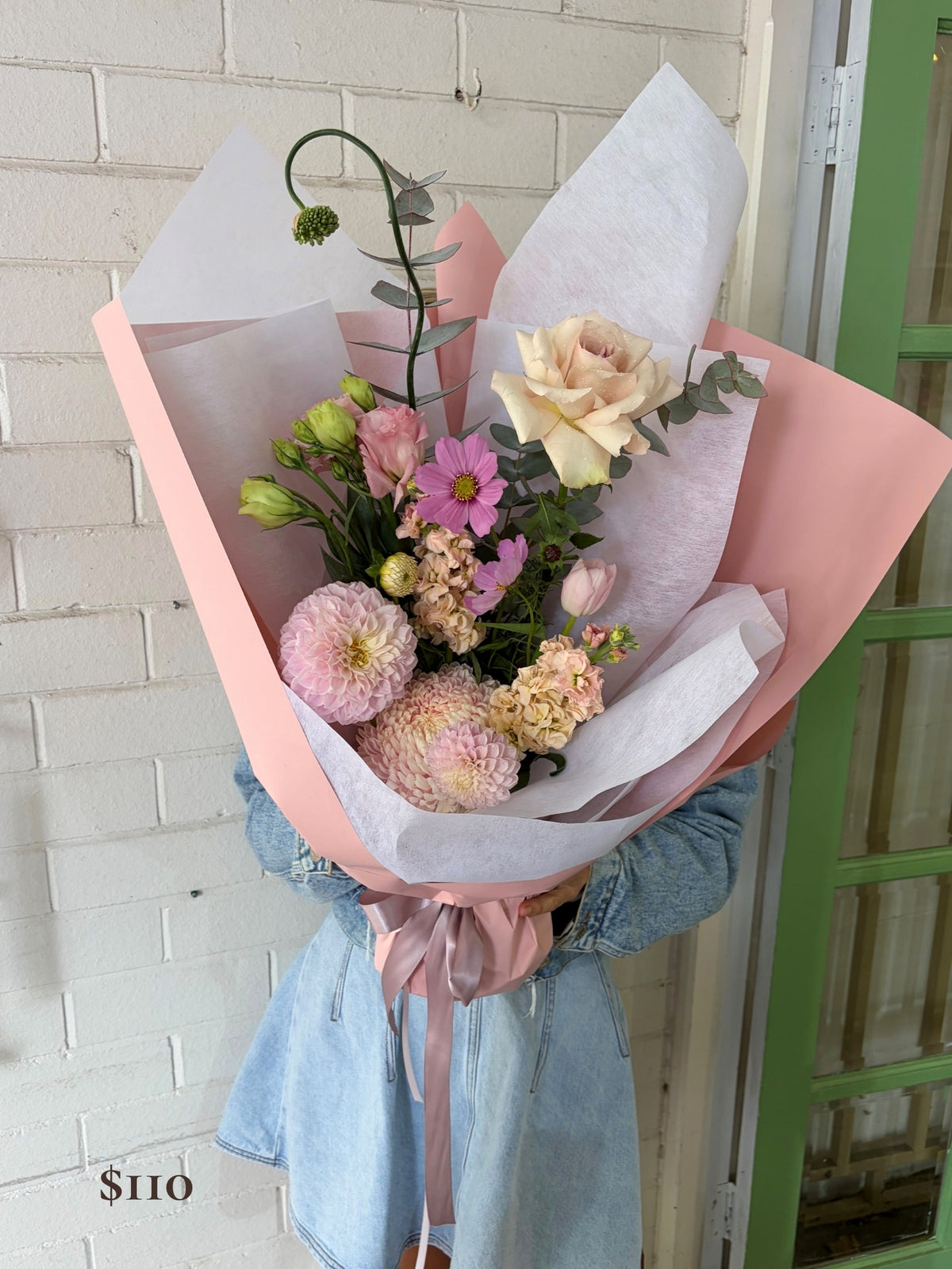 A hand-held bouquet of mixed wildflowers wrapped in pink paper and white tissue paper, held by a person wearing a light blue denim jacket in front of a white brick wall.